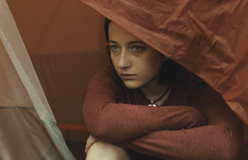 A young girl peaks out from beneath a tent flap, her arms folded over her raised knees.